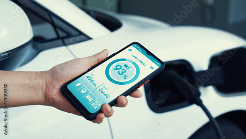Smartphone display battery status on smart EV mobile application while EV car recharging electricity by home charging station in garage. Future innovative EV car and energy sustainability. Peruse