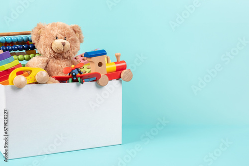 Toy box full of baby kid toys. Container with teddy bear, fluffy and educational wooden toys on light blue background. Cute toys collection for small children. Front view photo