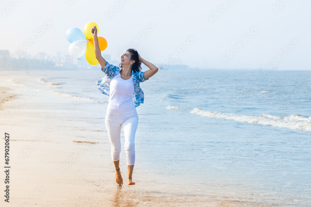 Beautiful young indian woman running with multicolor balloon in hands at the beach. Young girl having fun, holidays and vacation. - obrazy, fototapety, plakaty 