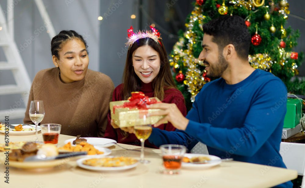 Asian cheerful female models wears reindeer antlers headband sitting smiling exciting when Idian male friend opening unboxing present gift box on dinner table celebrating Christmas eve in dining room