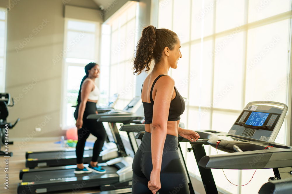 Fototapeta premium Fitness exercising in gym. Sporty woman in a sports bra doing exercise by running on treadmill during a workout at the gym fitness center with group of people.