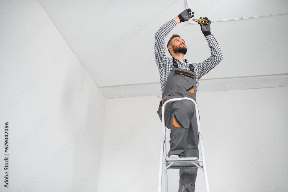 Repairman or professional electrician in workwear installing light spots, standing on the ladder in the white living room - obrazy, fototapety, plakaty 