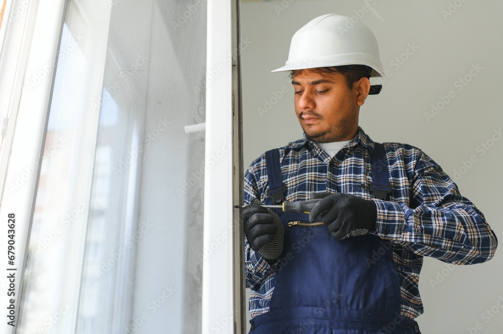 Indian Workman in overalls installing or adjusting plastic windows in the living room at home Indian Workman in overalls installing or adjusting plastic windows in the living room at home