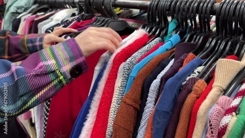 A young woman chooses clothes in a second-hand shop. Thrift store hangers with colorful sweaters. The concept of buying used clothes, fast fashion industry waste reduce and sustainability. Close-up.