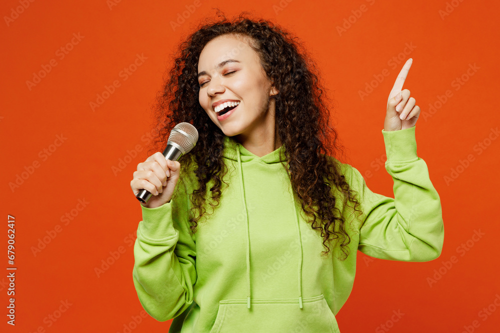 Young cheerful singer happy woman of African American ethnicity she wear green hoody casual clothes sing song in microphone at karaoke club isolated on plain red orange background. Lifestyle concept. Young cheerful singer happy woman of African American ethnicity she wear green hoody casual clothes sing song in microphone at karaoke club isolated on plain red orange background. Lifestyle concept.
