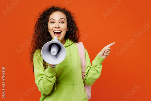 Young teen girl student of African American ethnicity wear casual clothes backpack bag hold books scream in megaphone point aside isolated on plain orange red background. High school college concept. photo