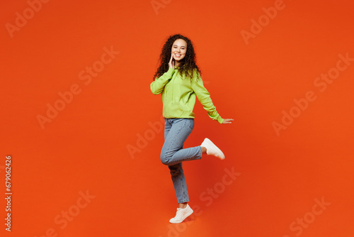 Fototapeta Naklejka Na Ścianę i Meble -  Full body side view nice happy young woman of African American ethnicity she wear green hoody casual clothes put hand on face raise up leg isolated on plain red orange background. Lifestyle concept.