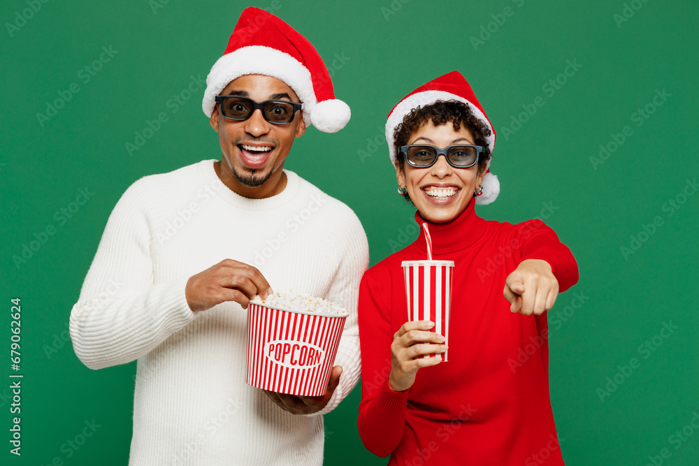 Φωτογραφία Merry surprised young couple friends man woman wear red casual clothes Santa hat posing 3d glasses watch movie film hold bucket of popcorn cup of soda pop in cinema isolated on plain green background