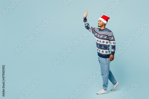 Full body side view young man wear knitted sweater Santa hat posing walk go strolling waving hand isolated on plain pastel blue background. Happy New Year 2024 celebration Christmas holiday concept.