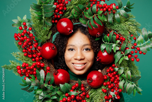 Close up smiling cheerful merry little kid teen girl posing hold looking camera through Christmas handmade wreath isolated on plain green background studio. Happy New Year celebration holiday concept.
