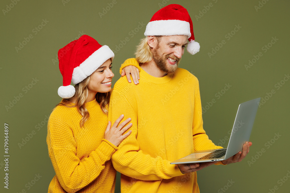 Merry young couple two IT friends man woman wear sweater Santa hat posing hold use work on laptop pc computer isolated on plain green background. Happy New Year celebration Christmas holiday concept. Merry young couple two IT friends man woman wear sweater Santa hat posing hold use work on laptop pc computer isolated on plain green background. Happy New Year celebration Christmas holiday concept.