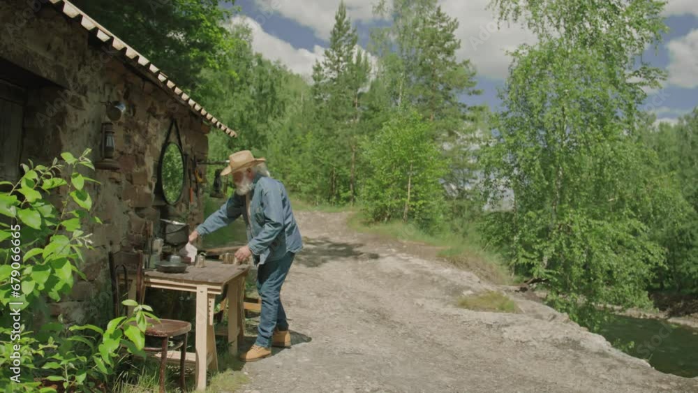 Very long shot of mature person standing next to table outside house at forest and organizing his workspace Very long shot of mature person standing next to table outside house at forest and organizing his workspace