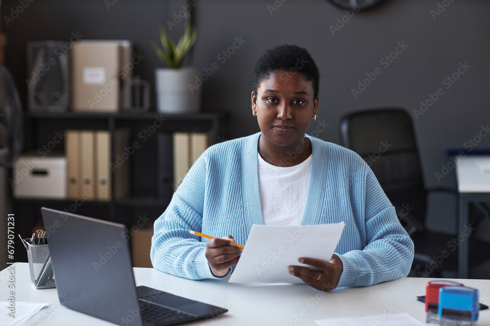 Young confident female manager of visa application center in casualwear looking at camera while sitting by workplace and checking data Young confident female manager of visa application center in casualwear looking at camera while sitting by workplace and checking data