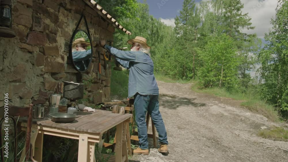 Long shot of elderly man using hammer while nailing tool hooks to facade of his house in forest Long shot of elderly man using hammer while nailing tool hooks to facade of his house in forest