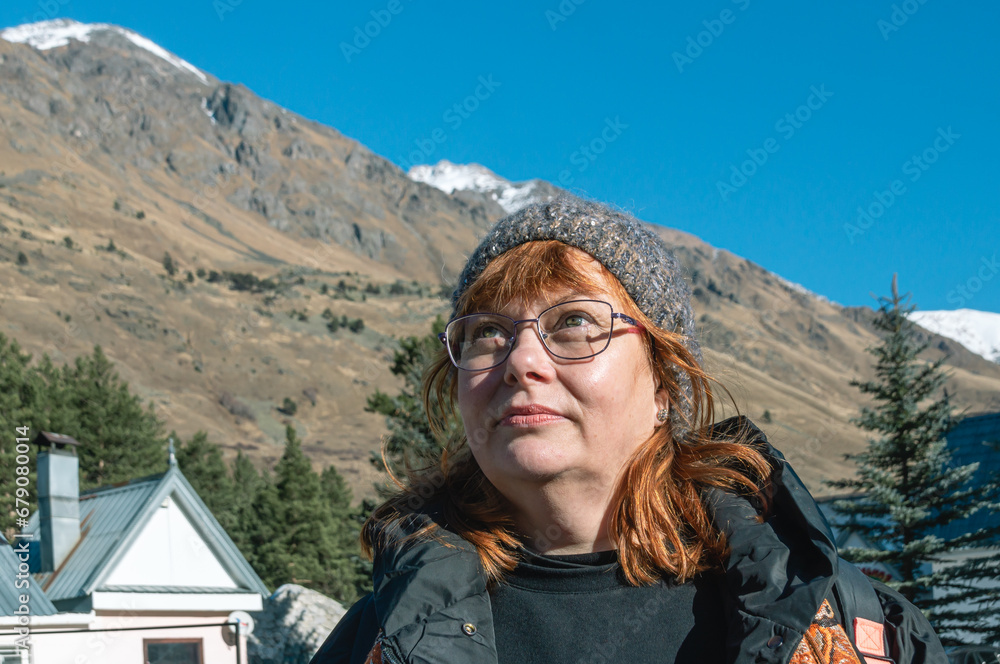 A woman with red hair and a warm knitted hat at a ski resort. Rest in the mountains. A woman with glasses looks at the mountains in bright sunny weather. Glasses for vision. A woman is in a good mood.