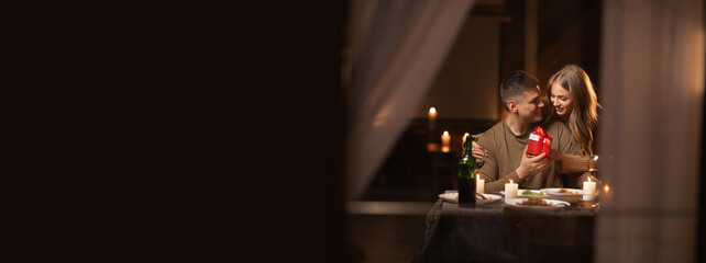 Young happy couple in love having a romantic candlelight dinner at home, husband giving wife a gift for anniversary or Valentine's Day