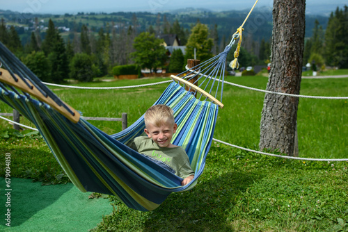 A little boy lies in a hammock in a recreation area against the backdrop of a beautiful mountain landscape. Summer vacation and vacation concept