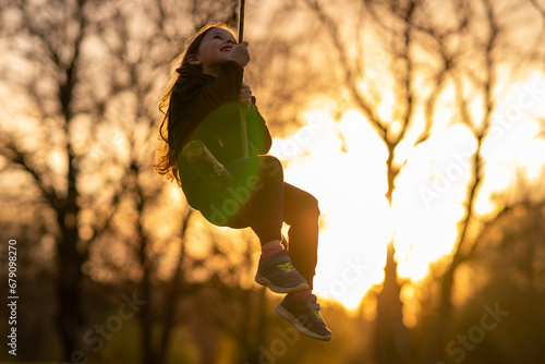 Happy childhood. Smiling little girl swings on homemade swing in nature, in village in sunset. Child protection.