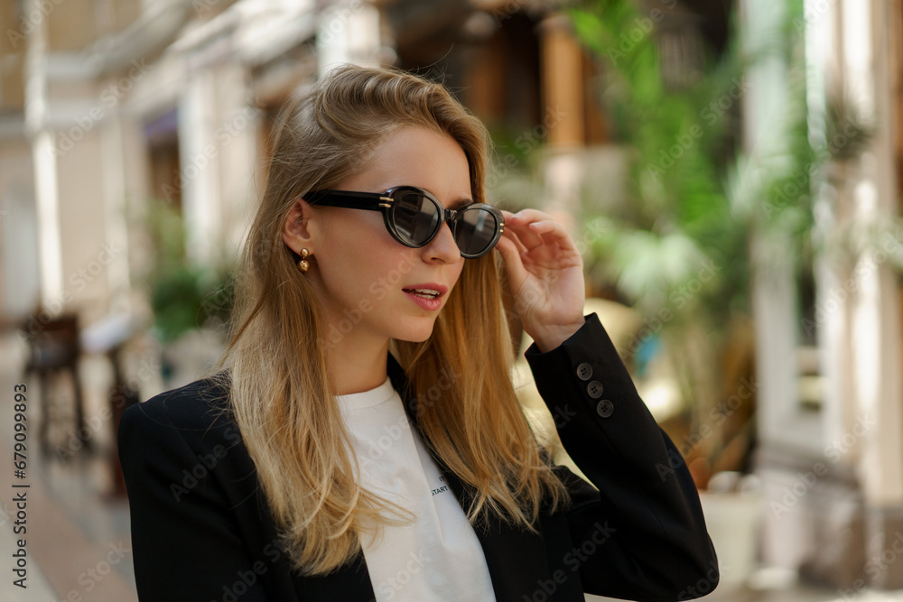 Close up portrait of pretty blond woman in stylish   casual business outfit walking  in  beautiful european city.  Wearing sunglasses.