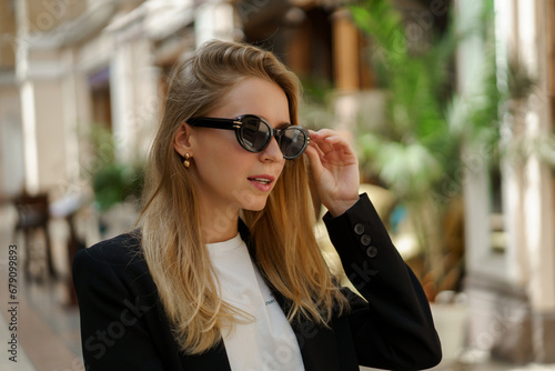 Close up portrait of pretty blond woman in stylish casual business outfit walking in beautiful european city. Wearing sunglasses.