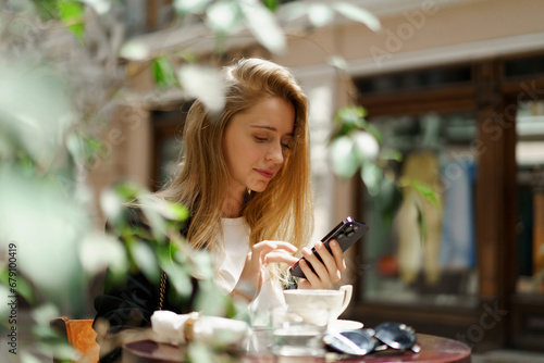 Beautiful blond woman in stylish casual business outfit sitting in cafe in beautiful european city. Using mobyle phone.
