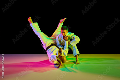 Two Caucasian karate fighters in white kimono with black belts performing skills in action in neon light isolated black. Concept of combat sport.