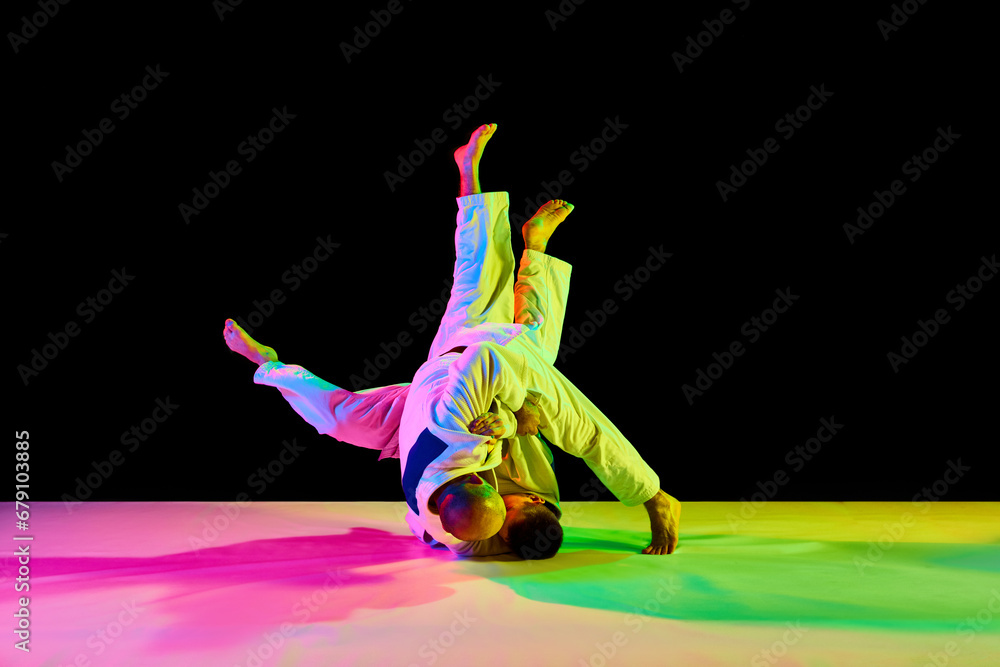 Two young, athletic professional karate men fighting, performing technical skill in neon light isolated black background.