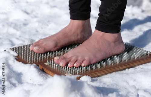 A woman practicing yoga therapy stands on a board with nails against a background of snow.