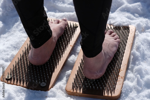 A woman practicing yoga therapy stands on a board with nails against a background of snow.