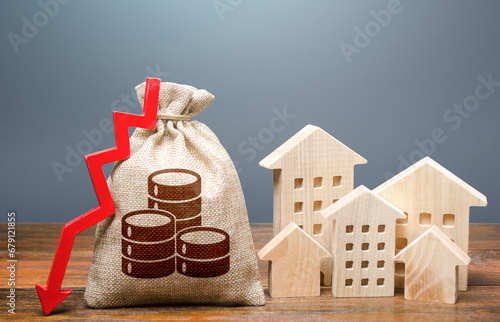 Money bag with red arrow down near wooden houses. Realty crisis. Falling of real estate market prices. Reducing maintaining cost, energy efficiency, saving. Low rates on mortgages. Taxes. Budget. photo