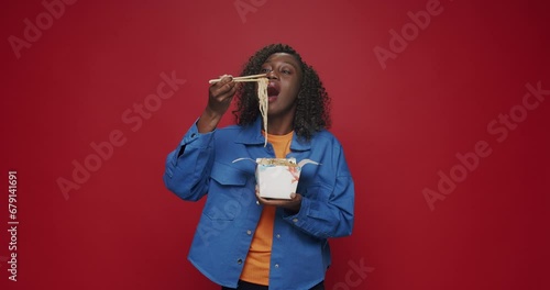 African american woman looking at camera and smiling, preparing to eat noodles, seafood and vegetables with chopsticks on isolated red background. Asia street food. Boxed fast food ready for delivery. photo