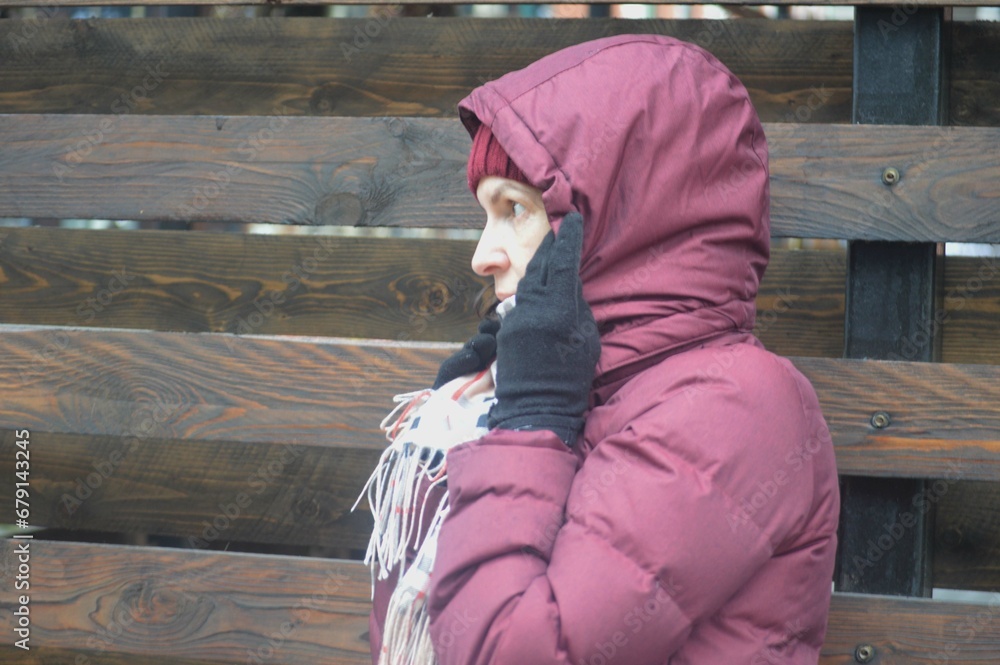 A Caucasian woman in a burgundy jacket and a checkered yellow scarf covers her head and face with a hood from the weather and wind. Weather protection. A Caucasian woman in a burgundy jacket and a checkered yellow scarf covers her head and face with a hood from the weather and wind. Weather protection.