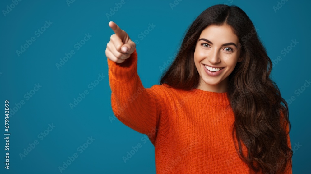 Fototapeta premium Portrait of a beautiful girl with long dark hair on a blue background wearing an orange sweatshirt, finger pointing in a side view