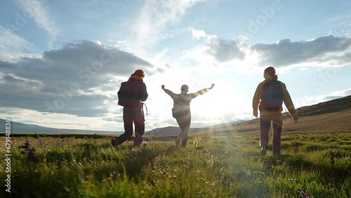 Three hiker women with backpacks walk along field at scenic mountain landscape in sun light. Active traveling way, tourist recreation and path in wild nature