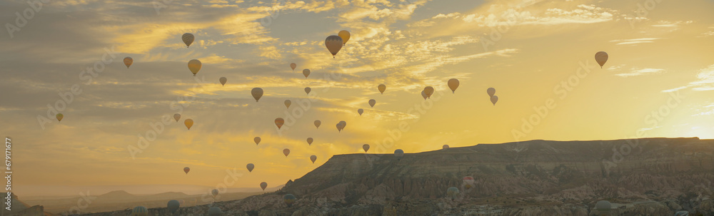 The valley with hot air balloons at sunrise amid the rocks, the sun rising from behind the mountains painted the clouds in golden color, Cappadocia, Turkey. - obrazy, fototapety, plakaty 