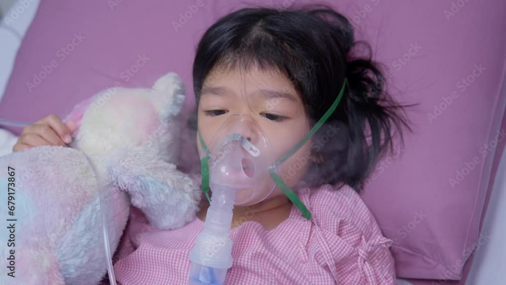 A little Asian girl has an oxygen mask and breathing through a nebulizer at the hospital. Concept of bronchitis, respiratory and Medical treatment, inhaling medicine, mist, from a nebulizer. A little Asian girl has an oxygen mask and breathing through a nebulizer at the hospital. Concept of bronchitis, respiratory and Medical treatment, inhaling medicine, mist, from a nebulizer.