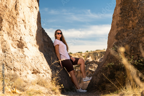 A young woman posing between rocks, leaning against one. Canyon crevice in the background. Sunny weather.