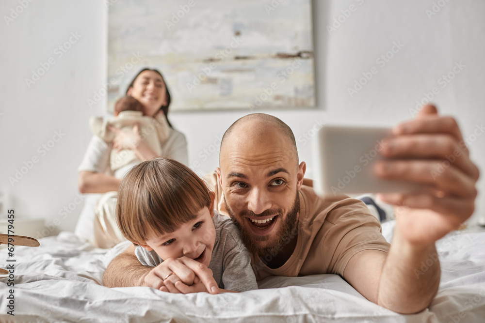 focus on cheerful father taking selfie with his son and his blurred wife and newborn on backdrop