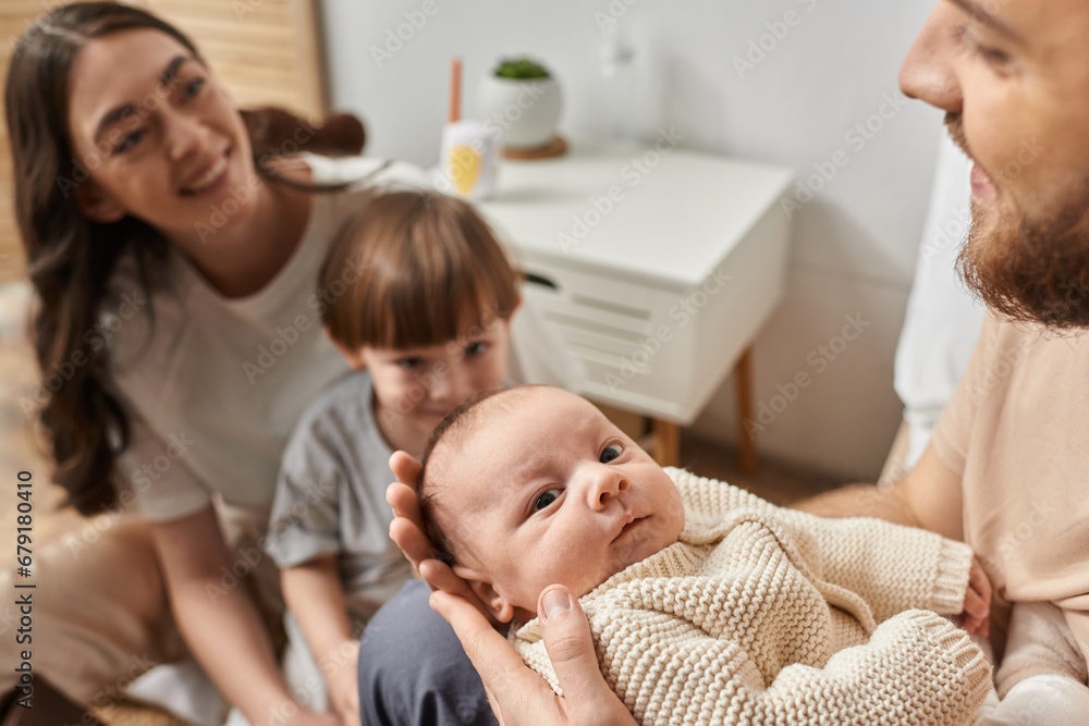 focus on cute newborn baby looking at camera surrounded by his blurred parents and brother, family focus on cute newborn baby looking at camera surrounded by his blurred parents and brother, family