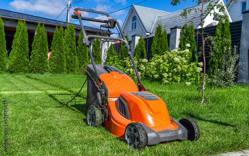 Electric lawn mower stands on the border of a mowed lawn and a luxuriantly growing grass in the backyard of country house at summer outdoors