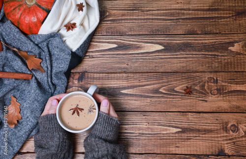 Cropped shot of female hands holding fresh coffee mug, latte, spicy cortado, bright orange pumpkin wrapped in woolen scarf, dry oak leaves, spices on rustic wooden table. Autumn background