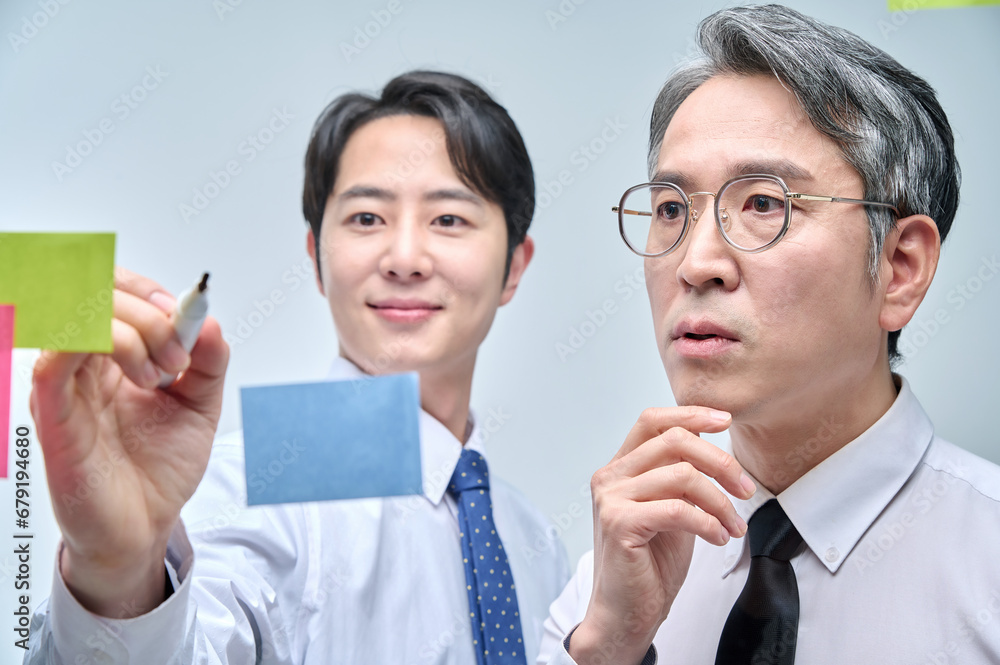 Two businessmen are standing on the glass wall of the office, looking at the attached paper and contemplating planning and strategy. Two businessmen are standing on the glass wall of the office, looking at the attached paper and contemplating planning and strategy.
