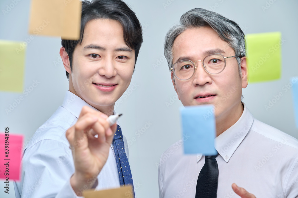 Two businessmen are standing on the glass wall of the office, looking at the attached paper and contemplating planning and strategy. Two businessmen are standing on the glass wall of the office, looking at the attached paper and contemplating planning and strategy.