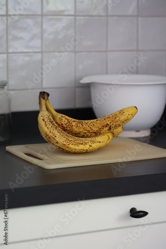 A portrait of a bunch of yellow bananas with brown spots lying on a white plastic cutting board on a black kitchen countertop. The delicious energizing food is ready to eat and ideal before sport.