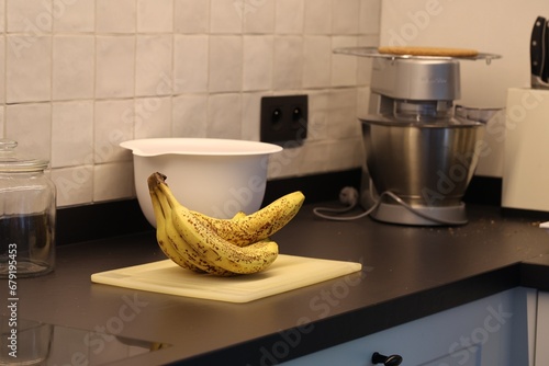 A portrait of a bunch of yellow bananas with brown spots lying on a white plastic cutting board on a black kitchen countertop. The delicious energizing food is ready to eat and ideal before sport.