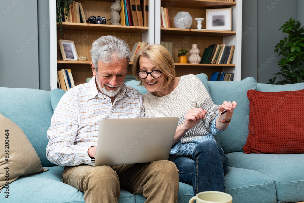 Cheerful old modern retired couple using credit card and laptop for online shopping, buying on Internet, choosing tour for vacation, making payment with bank application, financial service
