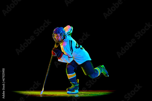 Photography Dynamic image of young girl, hockey player in uniform and helmet, in motion with stick against black studio background in neon light