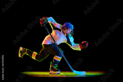 Competitive young woman, hockey player in motion, sliding on skates with pluck, training against black studio background in neon light. Concept of professional sport, competition, game, action, hobby