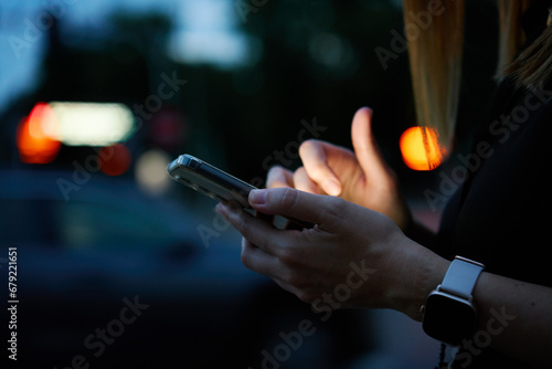 Close up shot of woman using smartphone on city street with bokeh lights at night. Mobile phone in female hands outdoors photo