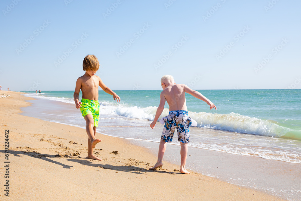 Two little boys playing on the beach on summer holidays. Joyful children in nature with beautiful sea, sand and blue sky. Happy kids on vacations at seaside running in the water and having fun